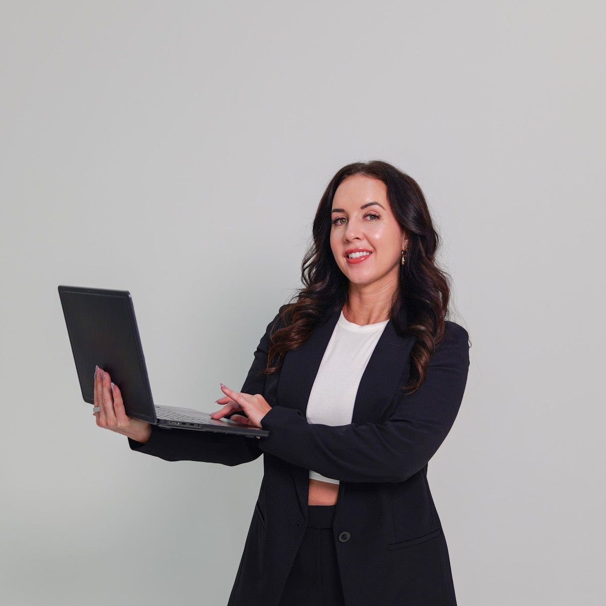 A woman in a black business suit holding a black laptop, standing against a solid light gray background.